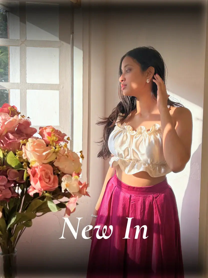 Woman wearing white ruffle crop top with maroon satin skirt, standing by window in soft sunlight, elegant new fashion collection