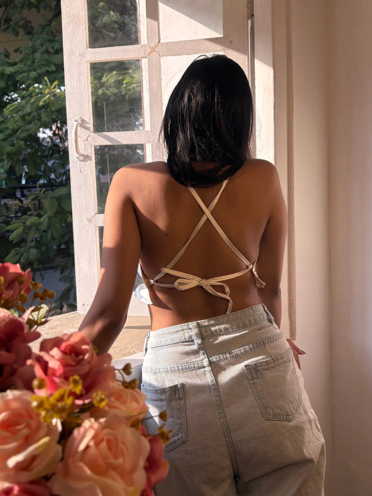 Person wearing a strappy top and light pants standing by a window with flowers in the foreground.