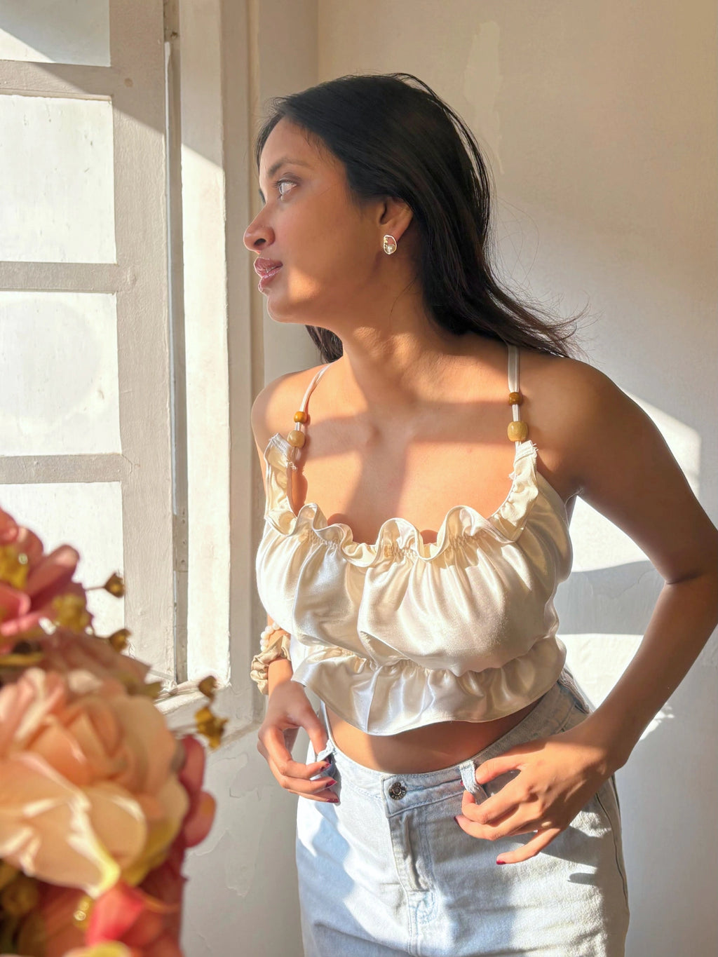 Woman wearing a white top with ruffled details and light blue jeans, standing indoors with a window and flowers in the background.