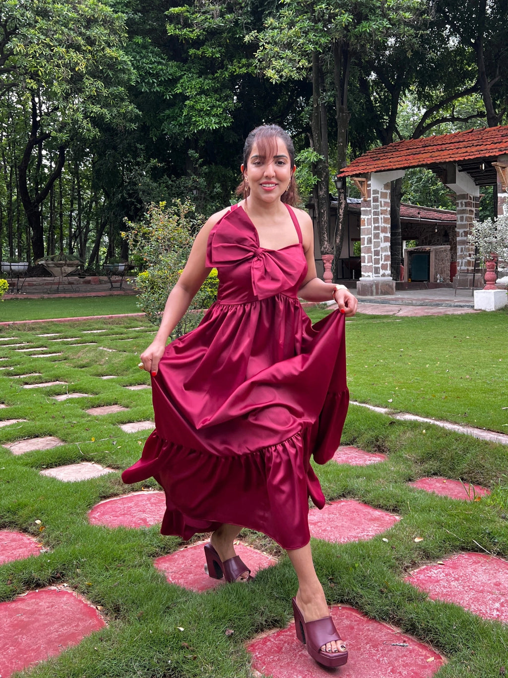 Woman in a red dress standing on a garden path with greenery and a building in the background