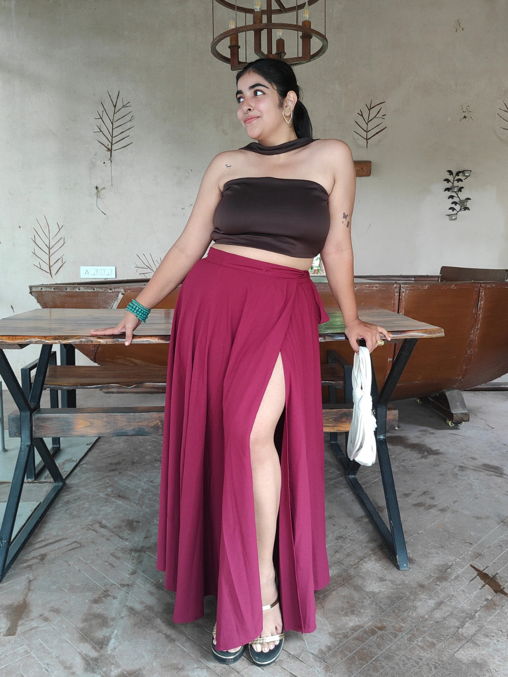 Woman in a brown top and maroon skirt standing in a casual setting with tables and chairs.