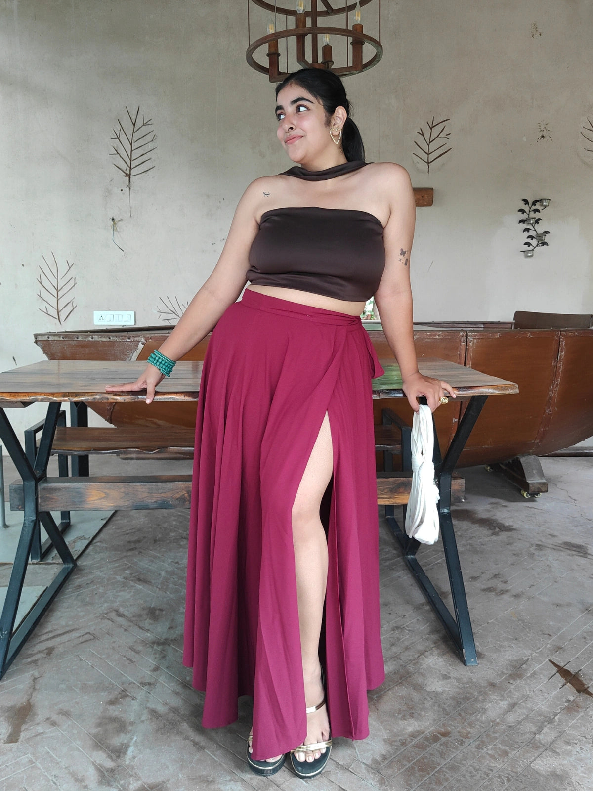 Woman in a brown top and maroon skirt standing in a casual setting with tables and chairs.