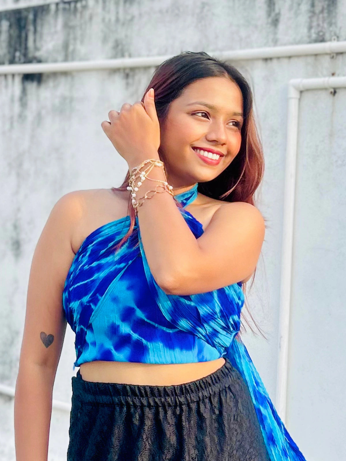 Woman wearing a blue tie-dye top and black pants, standing against a white wall.