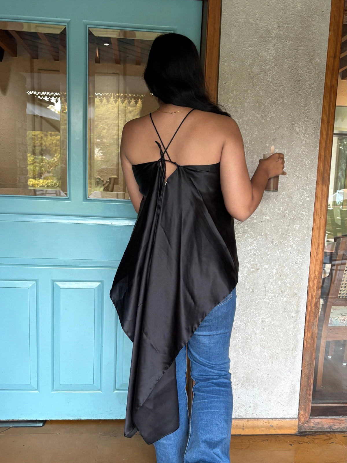 Person wearing a black satin top and blue jeans standing in front of a teal door.