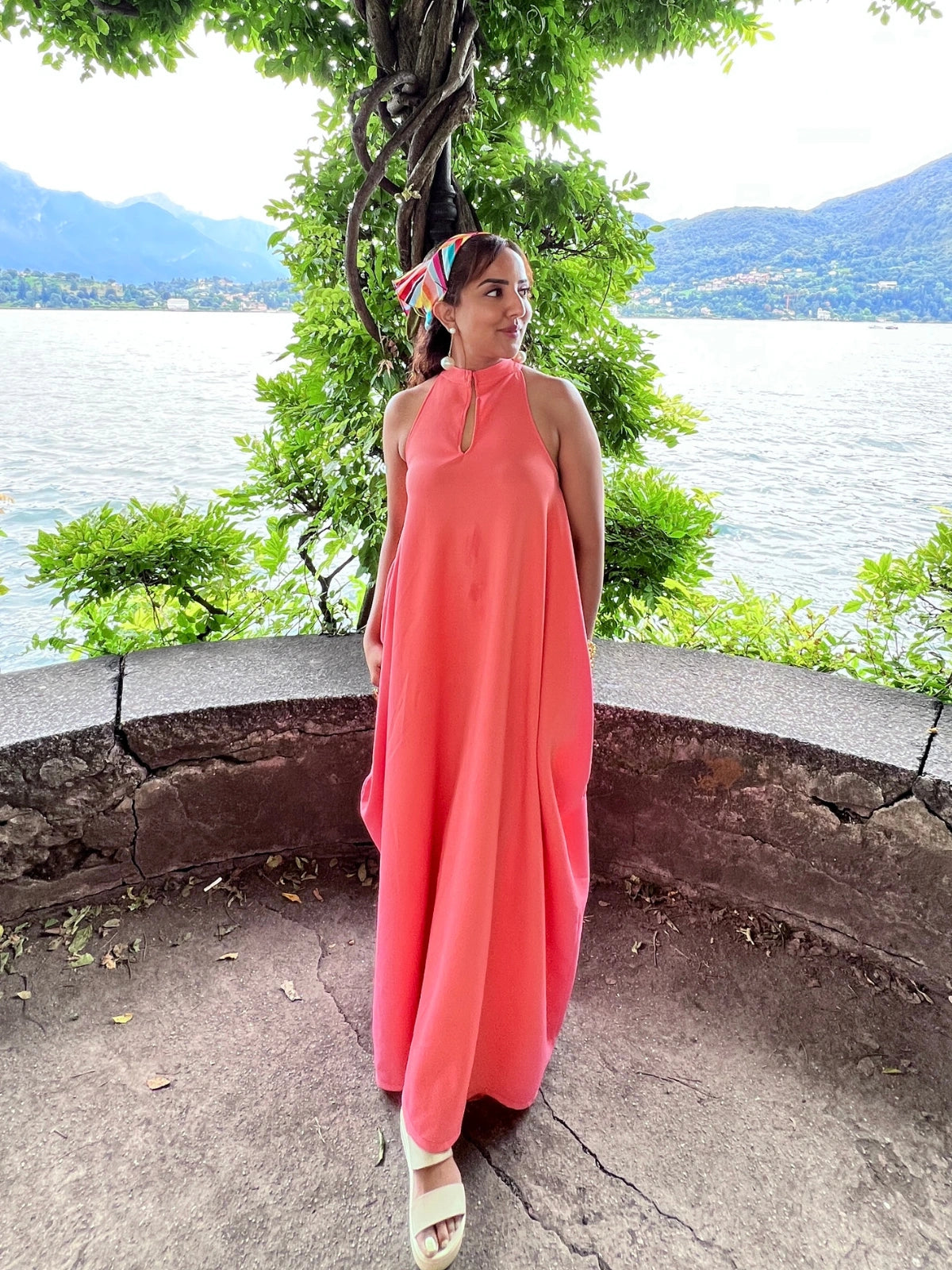 Woman in a pink dress standing by a scenic lakeside with mountains in the background.